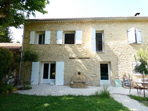 Un bâtiment en pierre doté de nombreuses fenêtres et portes, un petit jardin avec une table et des chaises, et un chemin en pierre menant à l’entrée.