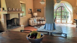 A cozy living room with a fireplace, a dining table with a fruit bowl, a sofa, and a large window with curtains.