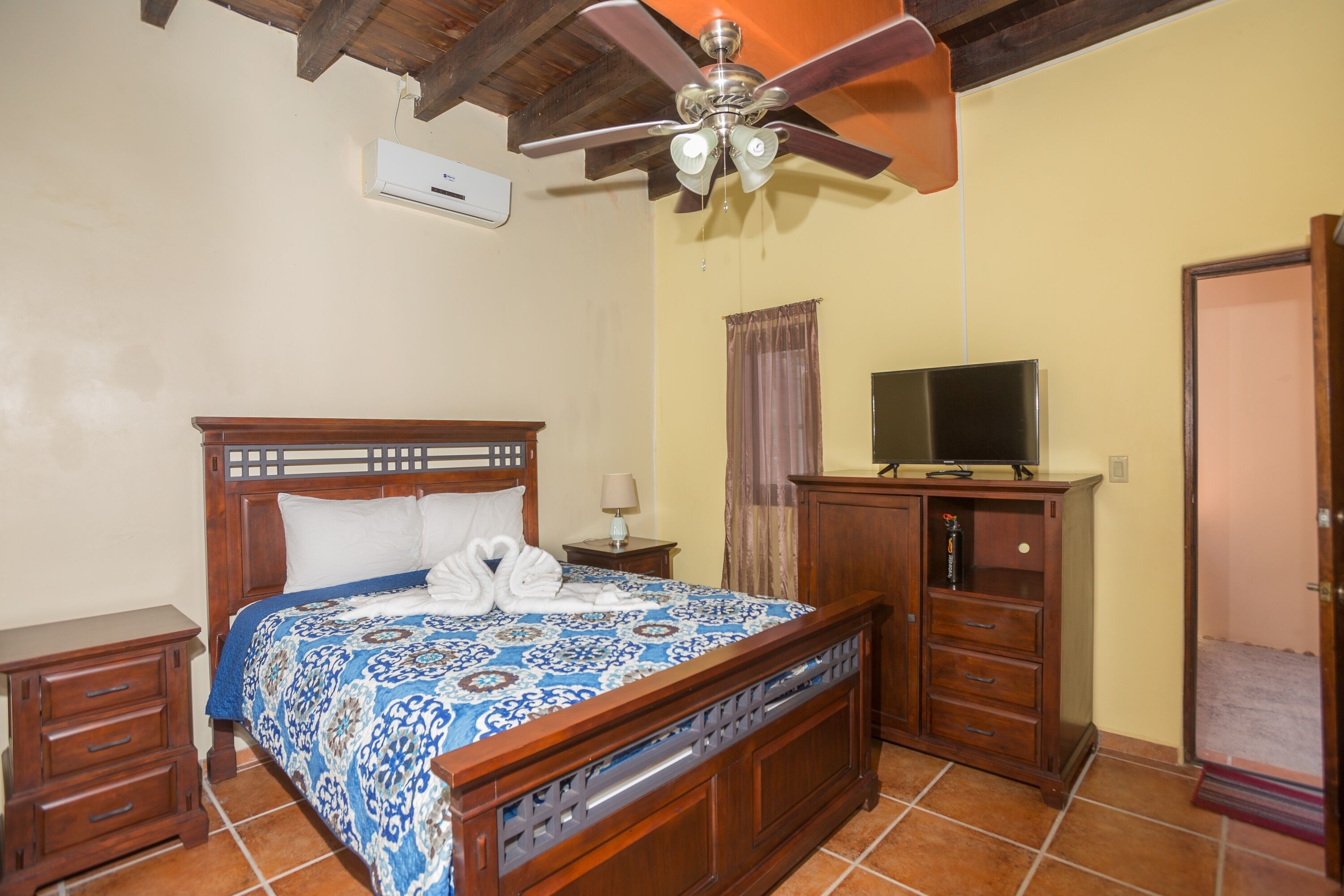 A bedroom with a wooden bed, a ceiling fan, a television on a wooden cabinet, and a door to the right.