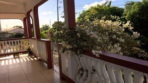 En balkon med tag, hvid gelænder, flisegulv og udsigt til grønt samt en bygning i baggrunden.