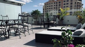 Una terraza en la azotea con mobiliario exterior, plantas en macetas y vistas de un edificio de varios pisos.