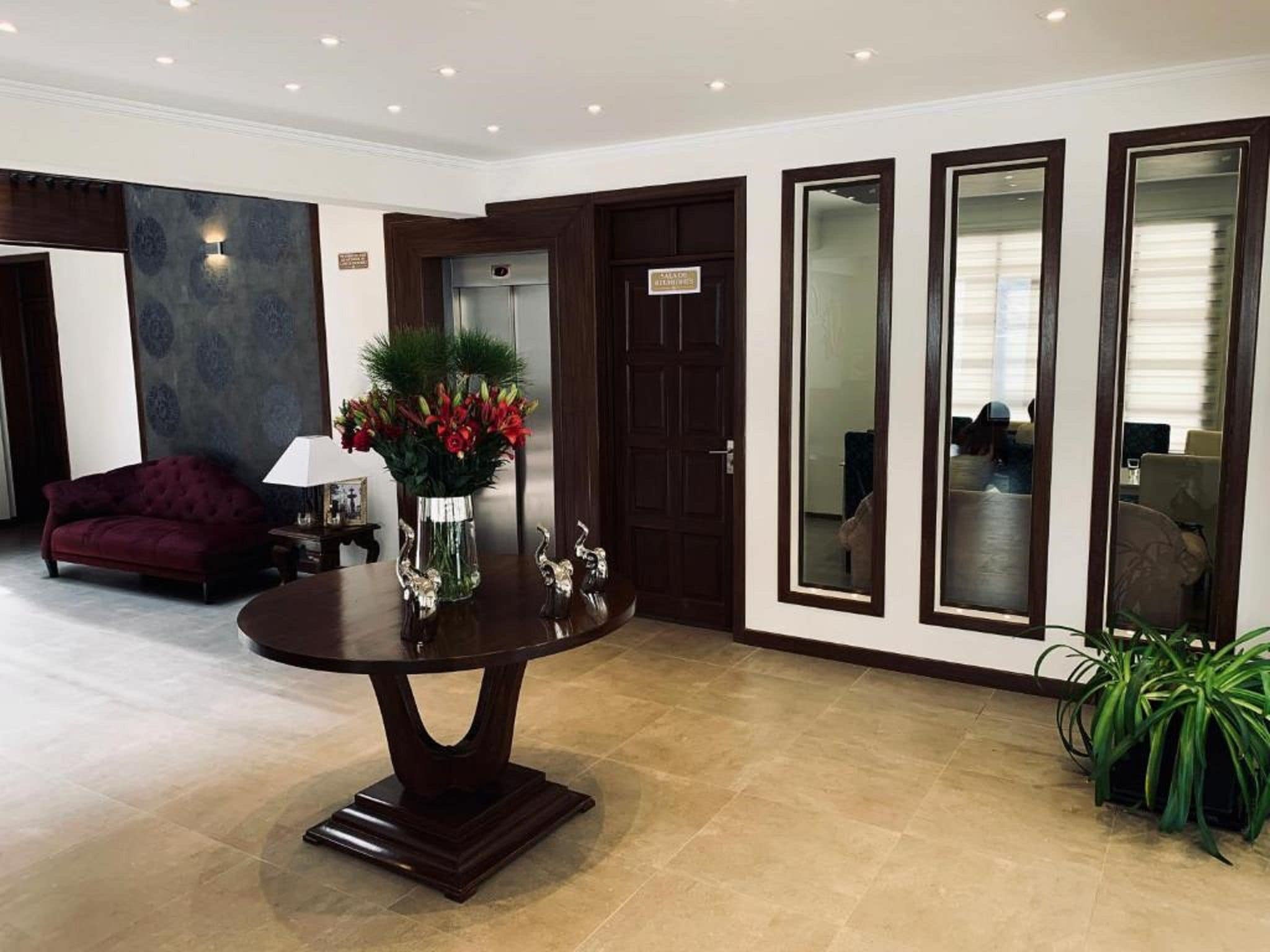 A lobby with a round table, a sofa, a vase with flowers, and a door with a sign.