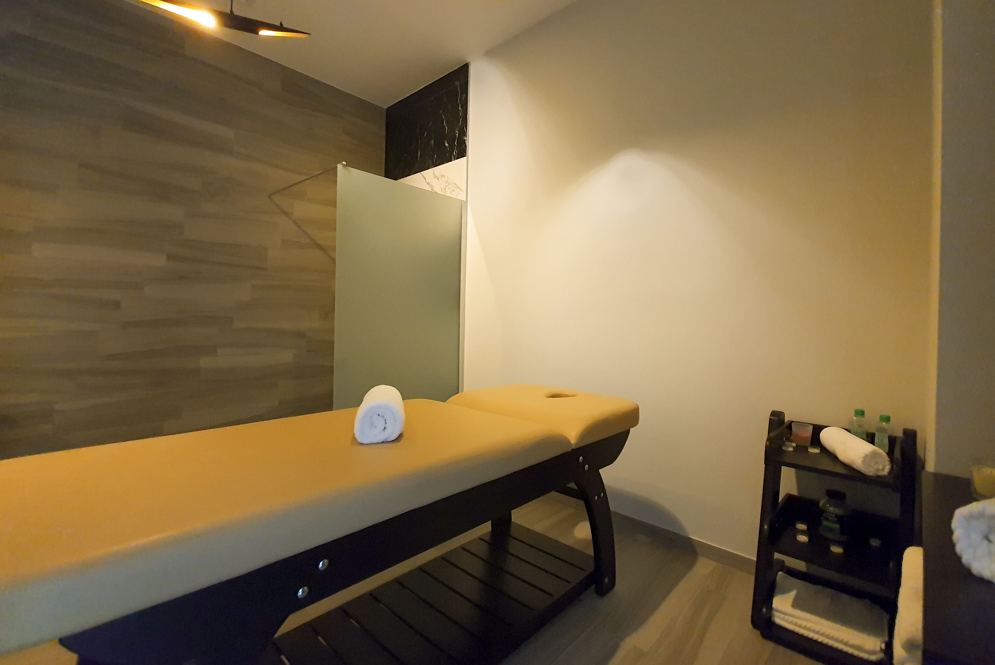 Ein moderner Massagetisch mit Handtuch und einer weißen Flasche, eine Holzwand, ein Spiegel und eine Ablage mit Handtüchern und Flaschen.