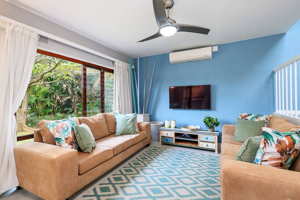Un salon avec un canapé beige, un tapis à motifs, un ventilateur de plafond et un téléviseur fixé au mur.