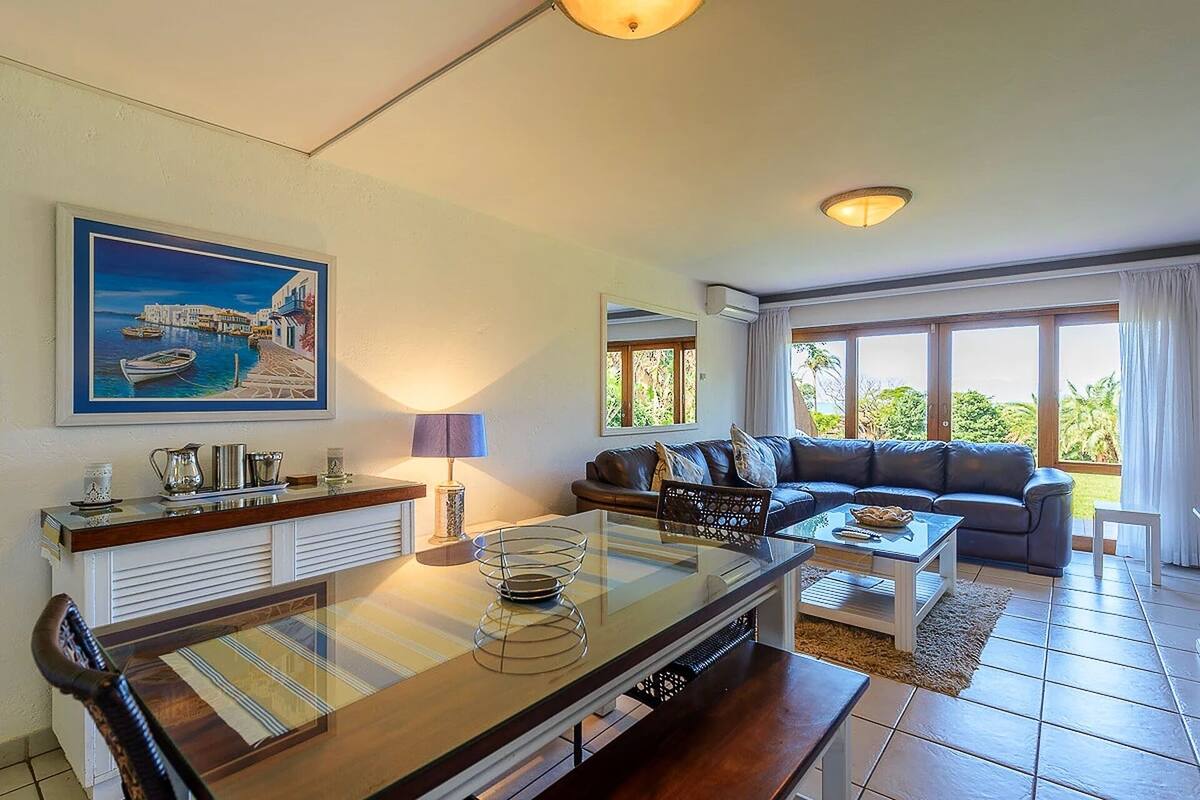 Un salon comprenant un canapé, une table basse en verre et un coin repas avec une table et des chaises. Un tableau est accroché au mur et il y a une fenêtre avec des rideaux.