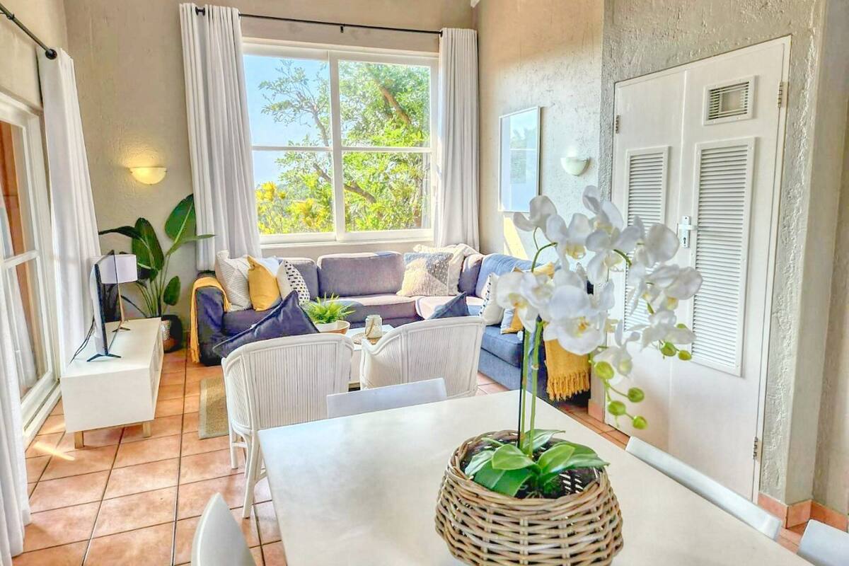 Un salon moderne avec un canapé blanc, une table à manger ornée d’une orchidée en pot et une grande fenêtre avec des rideaux.