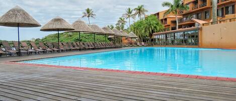 A swimming pool with lounge chairs and thatched umbrellas, surrounded by palm trees and a building with balconies.