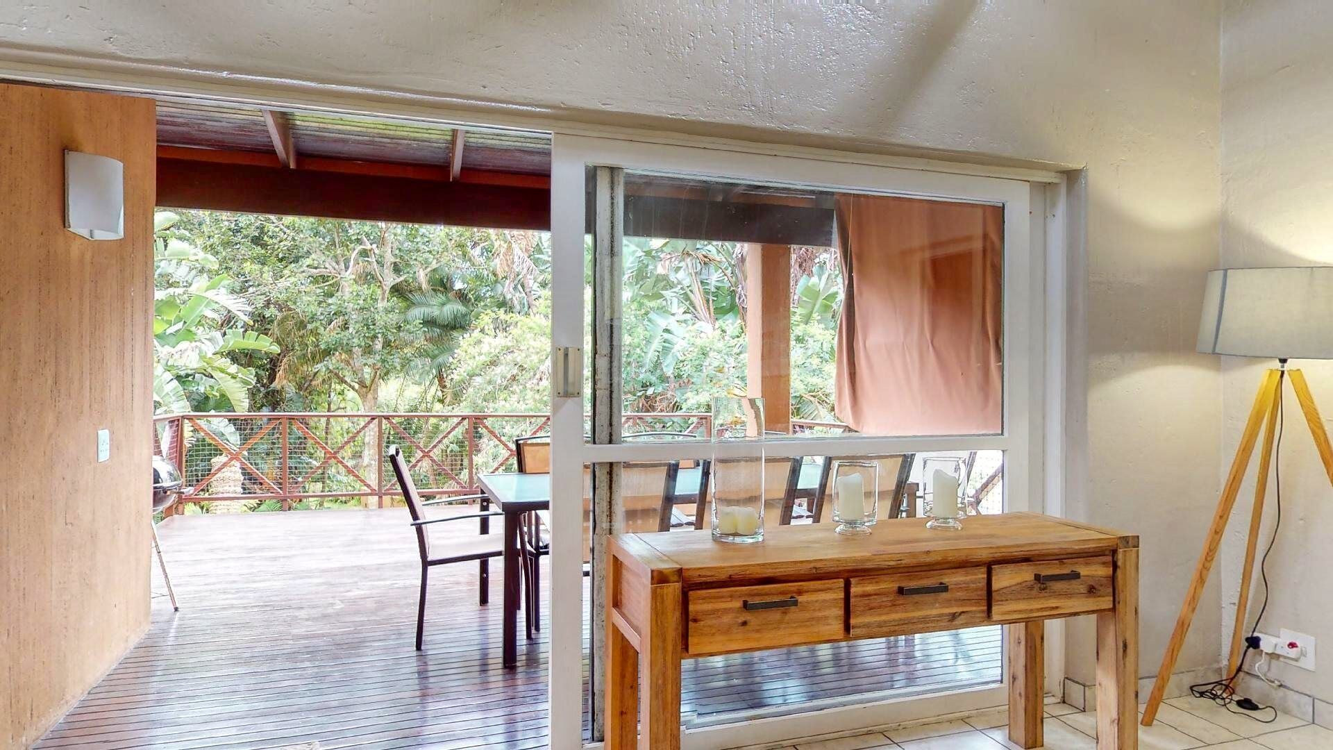 Una habitación con un escritorio de madera, dos sillas, un ventanal con vista a zonas verdes y una lámpara trípode.