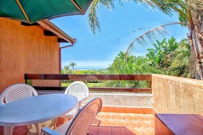Enjoy ocean views from this patio, featuring a round table and two chairs, shaded by a green umbrella amidst lush tropical surroundings.