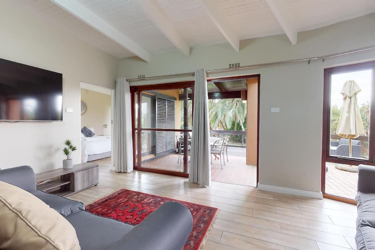 Un salon moderne avec un téléviseur à écran plat, un canapé, une table basse et un tapis rouge à motifs. Une porte-fenêtre coulissante donne accès à un patio extérieur aménagé avec une table et des chaises.