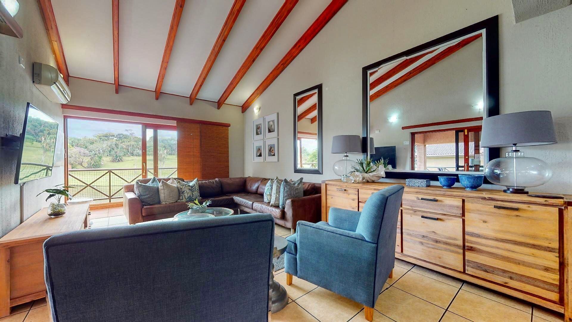A spacious living room with a large sofa, a coffee table, and a blue armchair. There is a wooden cabinet with a mirror and a lamp. The room has large windows with a view of greenery outside.