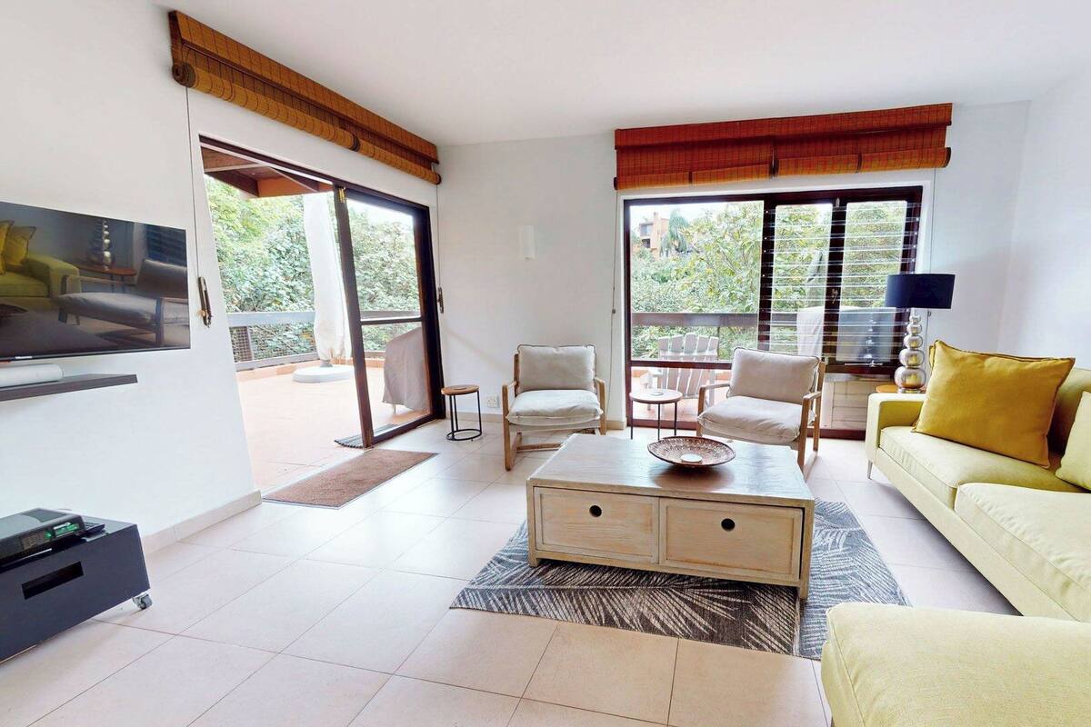 Un salon spacieux avec un canapé jaune, une table basse et deux fauteuils. On y trouve une télévision sur le côté gauche et un balcon avec vue sur la verdure.