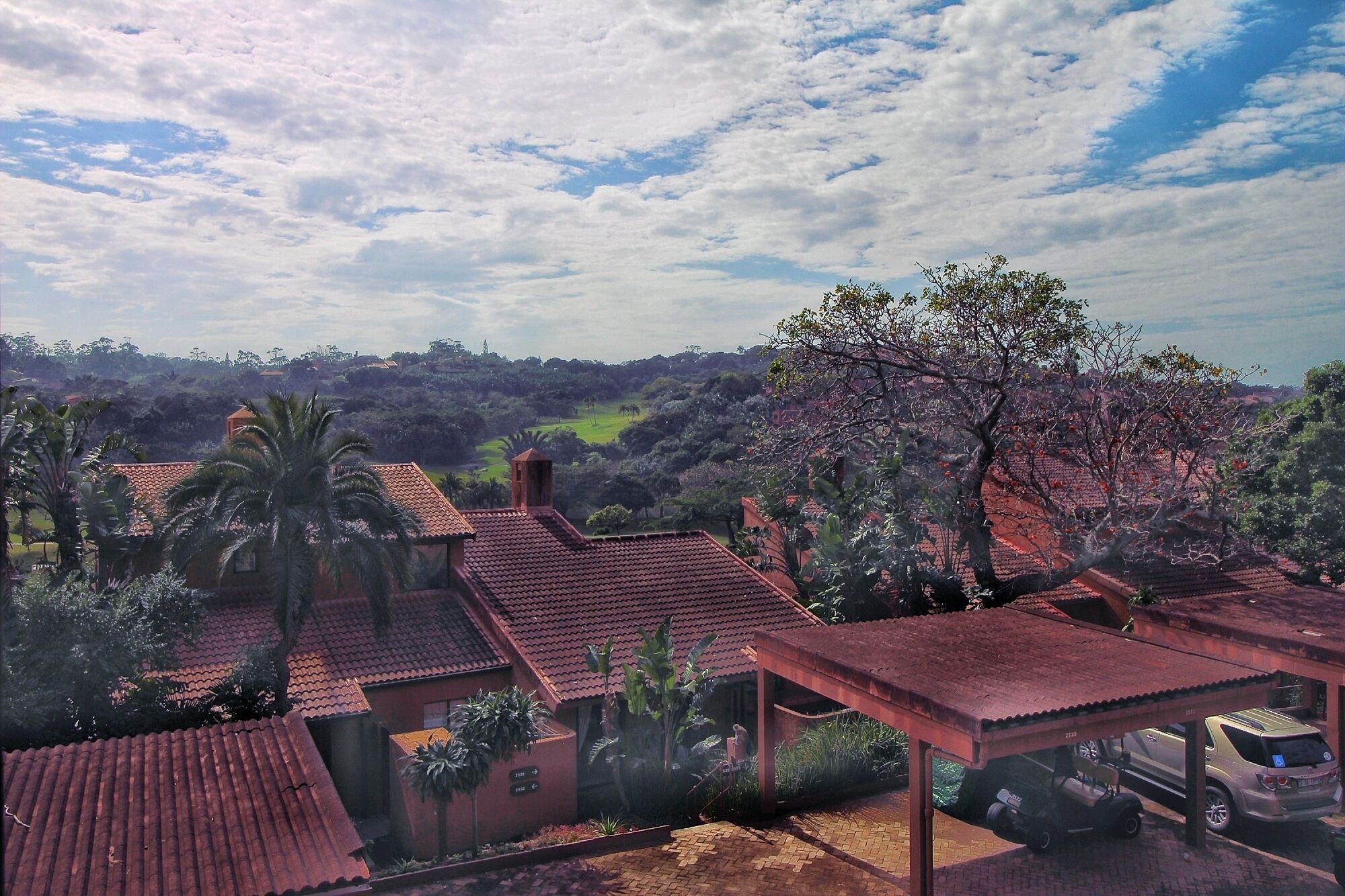 A residential area with houses featuring red-tiled roofs, surrounded by lush greenery and trees.
