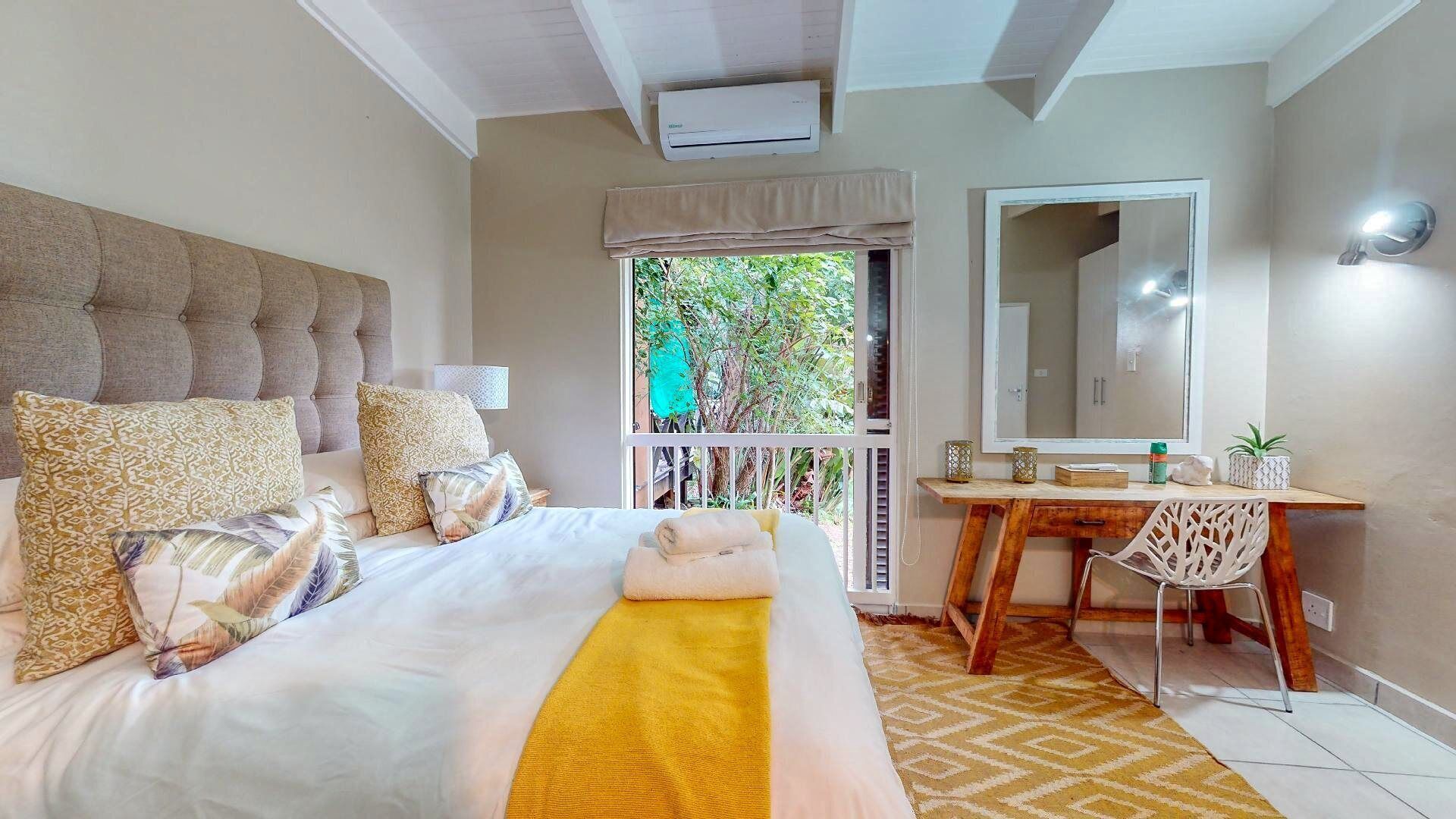 A bedroom with a large bed, a desk, a chair, and a window with a view of greenery.