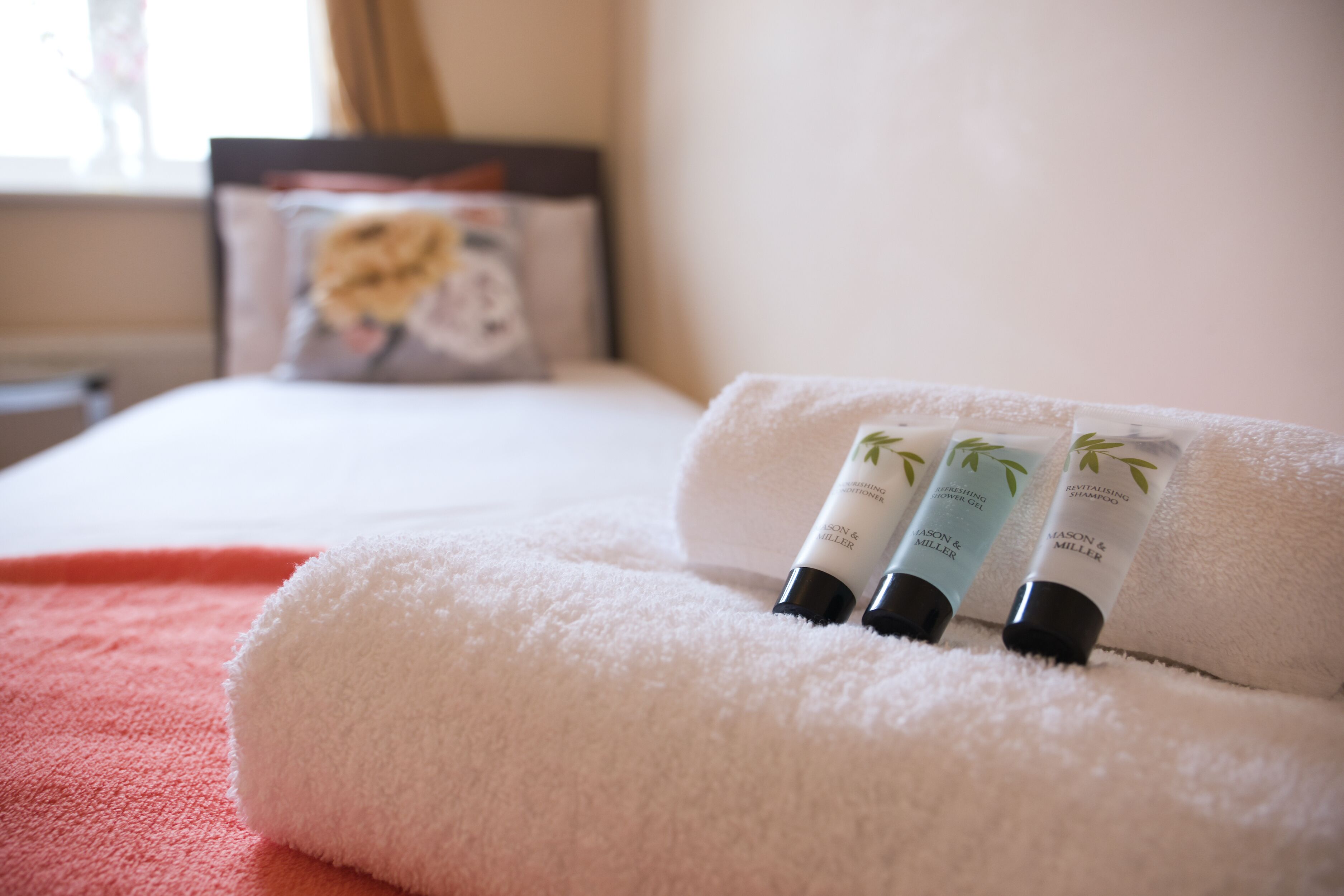 A neatly made bed with a white towel rolled up at the foot, and three bottles of L'Occitane shower gel placed on the towel.
