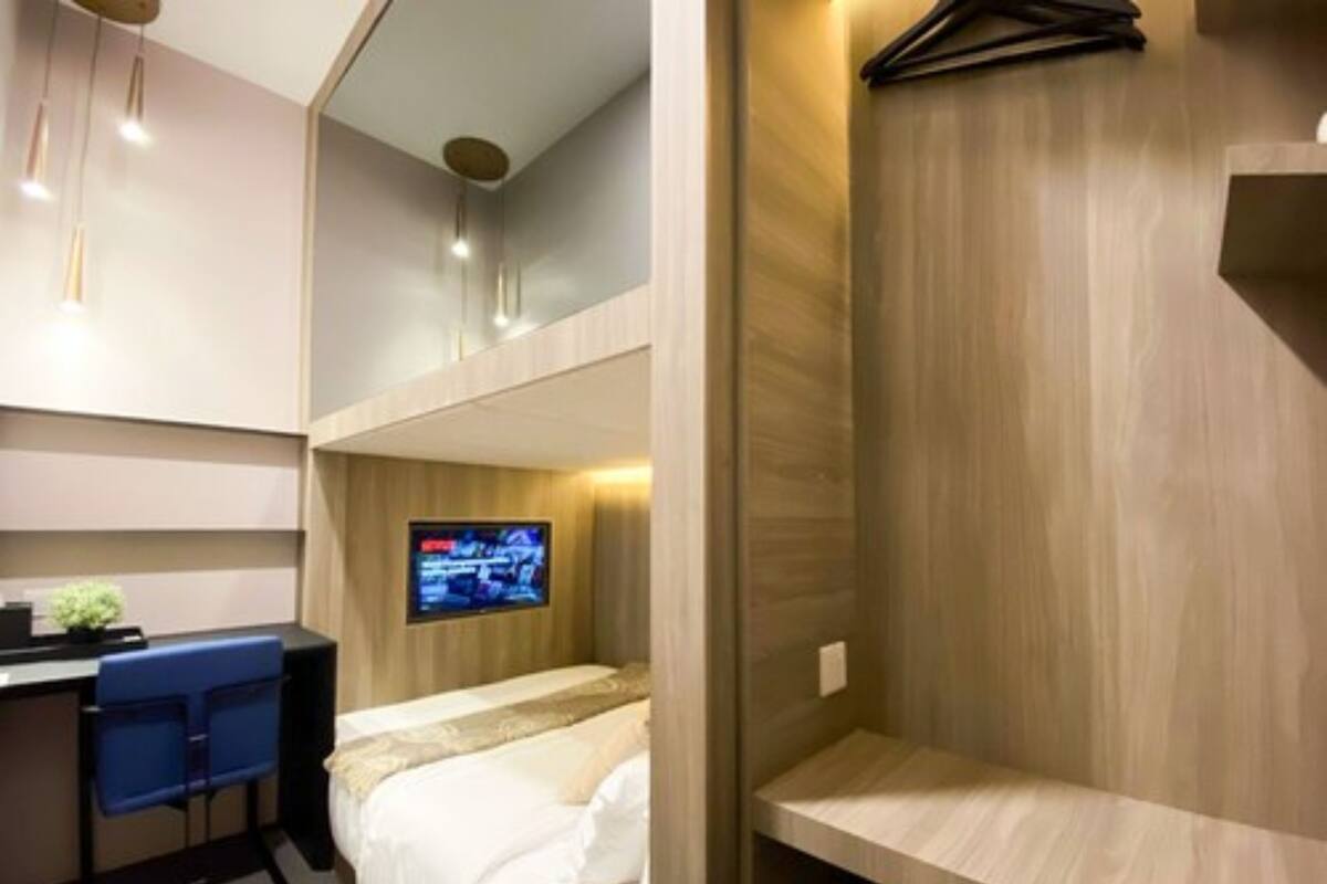 A compact hotel room with a bed, a desk with a chair, a TV mounted on the wall, and a wooden shelf.