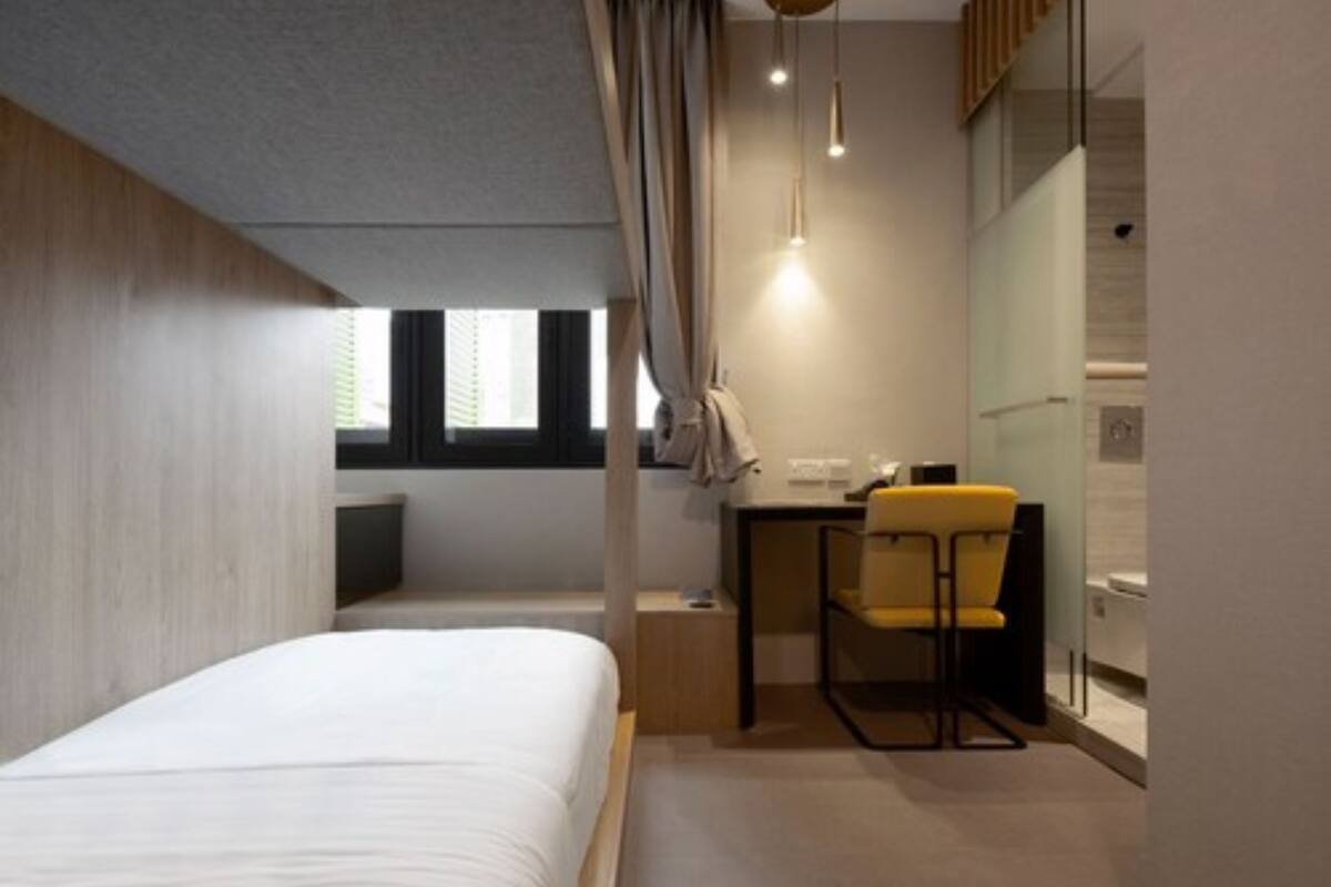 A modern hotel room with a bed, a desk, and a chair. There is a window providing natural light and a view of the outside.