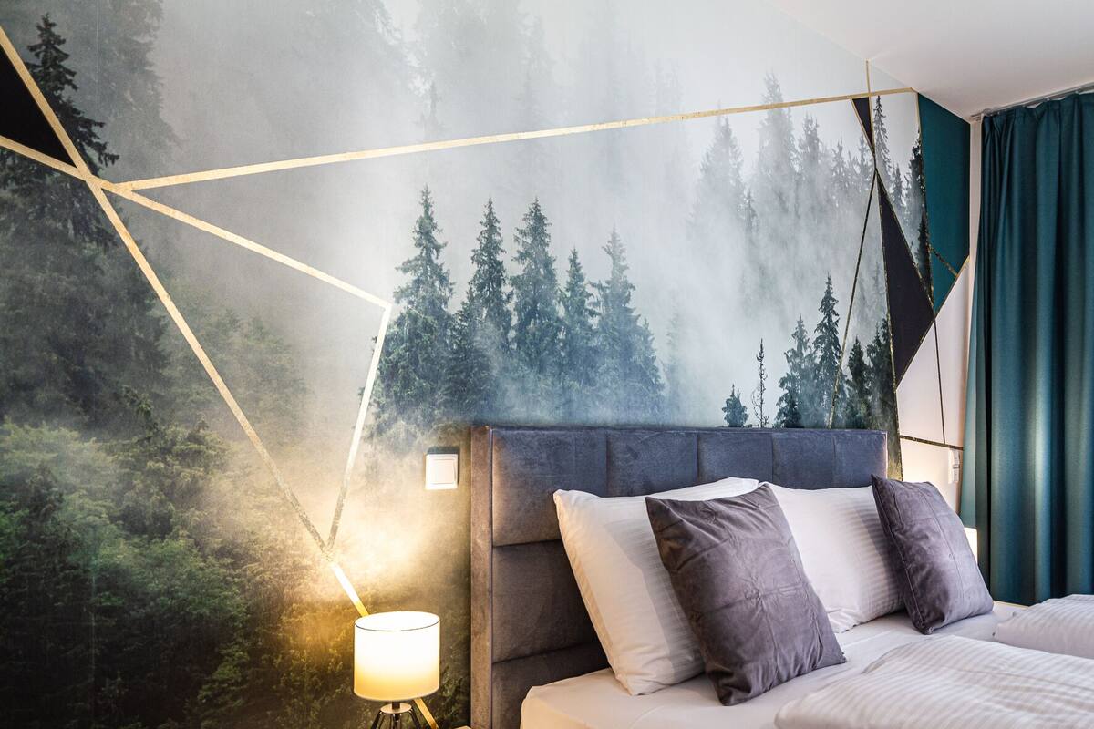 A bedroom with a bed, a nightstand, a lamp, and a wall mural of a forest.