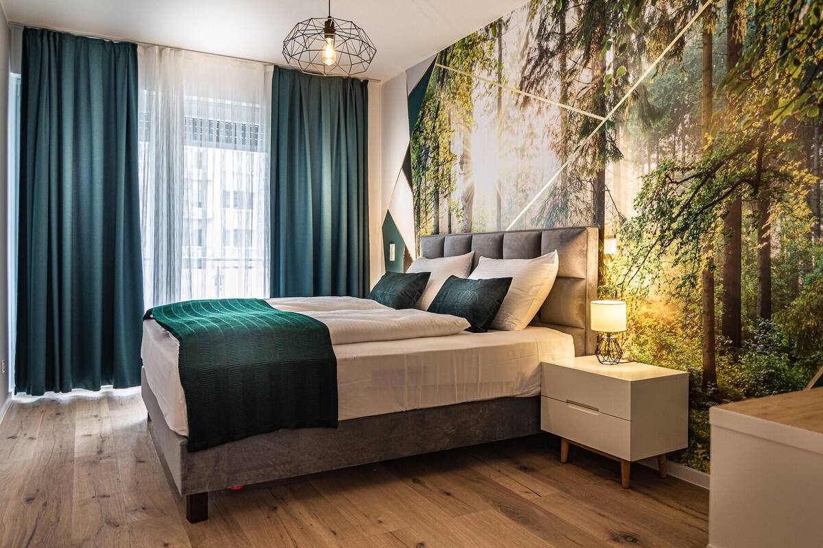 A modern bedroom with a large bed, a nightstand, and a wall mural of a forest.