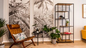 Uma sala de estar com uma cadeira de fibra natural, um aparador de metal preto e um grande mural com tema tropical.