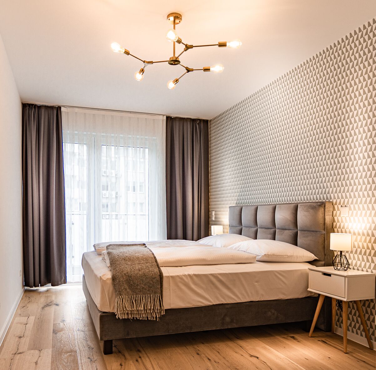 A modern bedroom with a large bed, a geometric patterned wall, a ceiling light fixture, and a nightstand with a lamp.