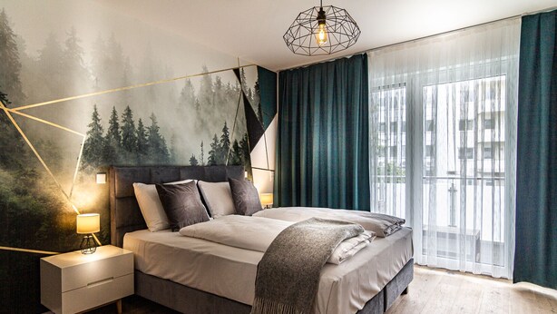 Quarto moderno com uma cama grande, mesa de cabeceira com abajur, luminária geométrica e um mural com tema de floresta na parede.