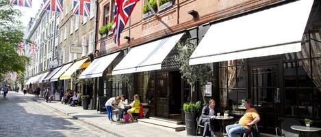 A row of outdoor seating areas with tables and chairs, a brick building with a sign reading "HOSPITAL," and multiple British flags.