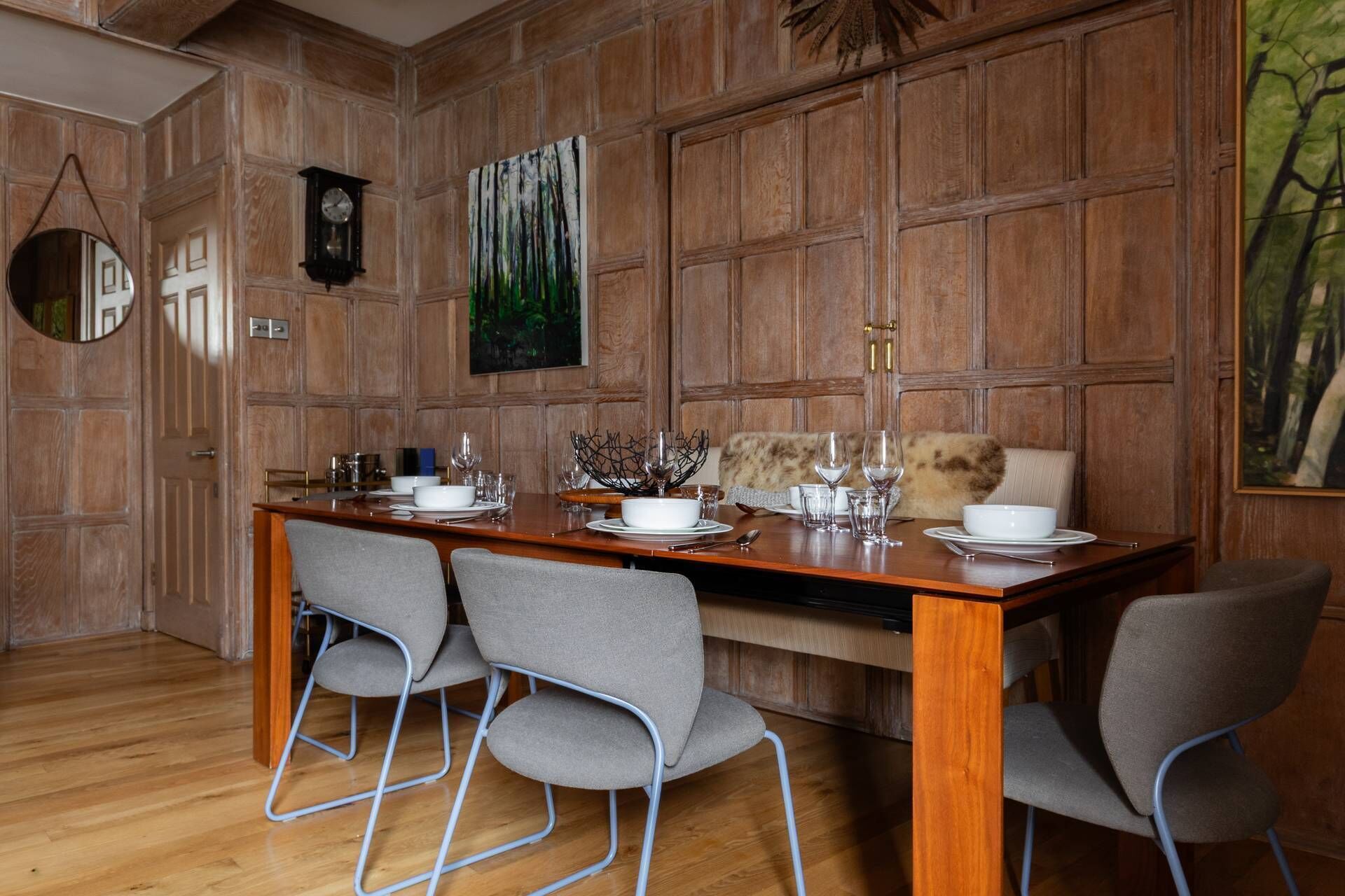 Un comedor con una mesa de madera dispuesta para cuatro personas, sillas tapizadas de color gris, un cuadro de un bosque, un reloj y un espejo en la pared.