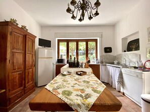 A kitchen with wooden cabinets, a white refrigerator, a dining table with a floral tablecloth, a chandelier, and a window with a view of greenery.