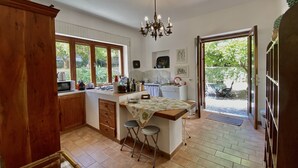 A kitchen with a wooden cabinet, a dining table with two stools, a chandelier, and a view of the garden through large windows. - Dimora Tana Pacenzia (Cefalù)