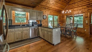 A wooden cabin with a kitchen featuring a refrigerator, dishwasher, and a dining area with a table and chairs. - Majestic Sunrise- Pet Friendly | Screened Porch | Hot Tub (Blue Ridge)