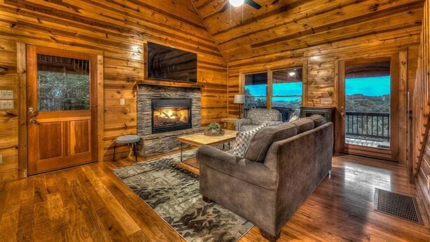 A log cabin interior with a stone fireplace, wooden flooring, a sofa, and a view of the mountains through the windows. - Majestic Sunrise- Pet Friendly | Screened Porch | Hot Tub (Blue Ridge)