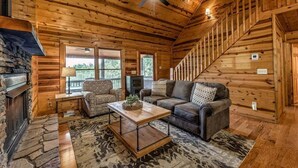 A cozy cabin interior with a fireplace, wooden walls, a sofa, a chair, and a staircase. - Majestic Sunrise- Pet Friendly | Screened Porch | Hot Tub (Blue Ridge)