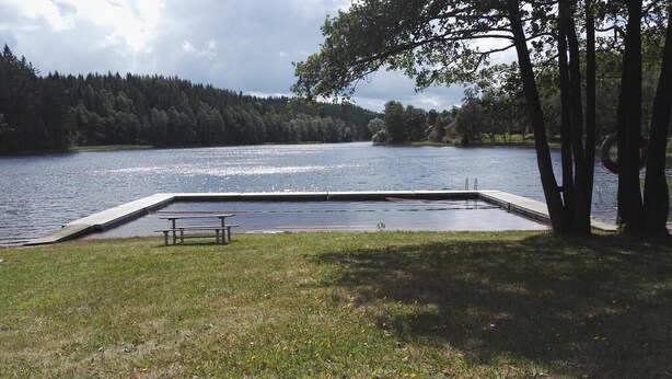 A swimming pool with a bench, surrounded by grass and trees, with a lake and forest in the background. - Lins B&B (Älvsered)