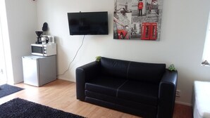 A living room with a black leather sofa, a flat-screen TV mounted on the wall, a small kitchenette with a microwave and coffee maker, and a poster of London on the wall. - Lins B&B (Älvsered)