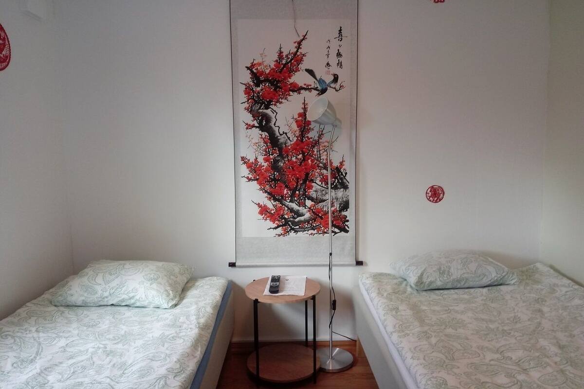 Ein Zimmer mit zwei Betten, einem kleinen Tisch mit Fernbedienung und einem an der Wand hängenden Bild von einem Baum mit roten Blüten und Vögeln.