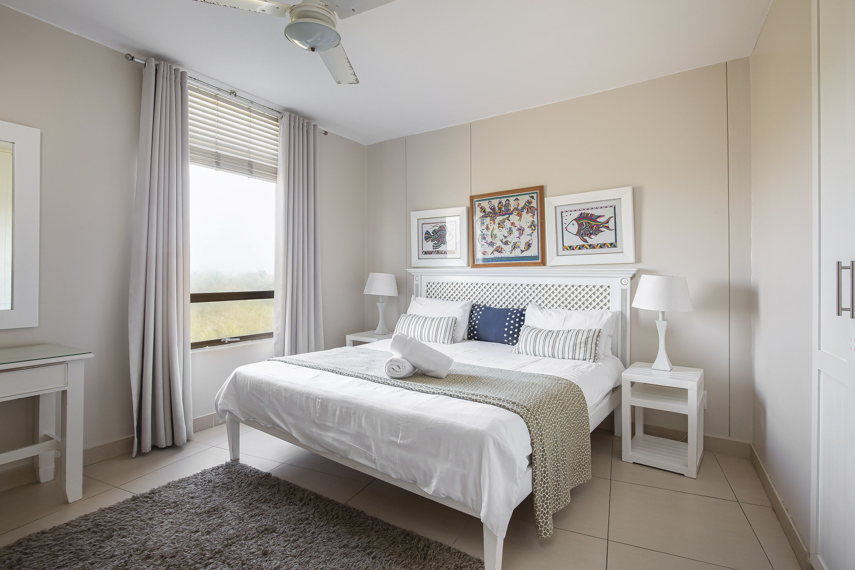 Un dormitorio ordenado con cama, mesitas de noche, escritorio y ventana con cortinas.