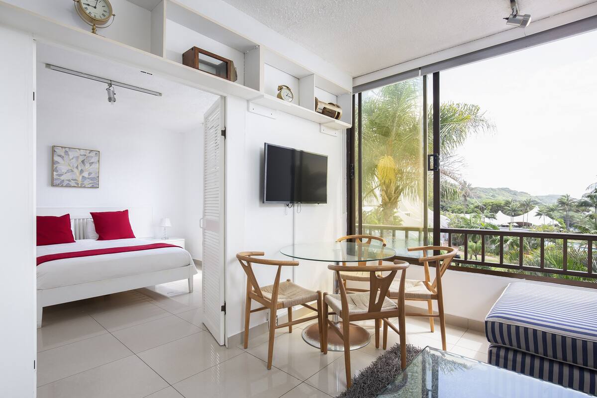 Een moderne slaapkamer met een bed, een televisie, een balkon met een tafel en stoelen, en uitzicht op groen.