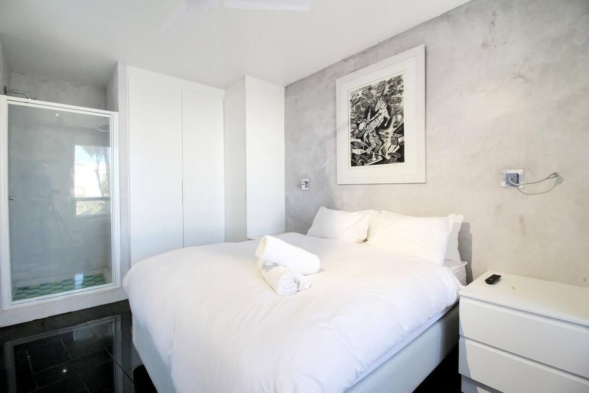 Een moderne hotelkamer met een groot bed, een witte nachtkastje, een kunstwerk in een lijst aan de muur en een douchecabine van glas.