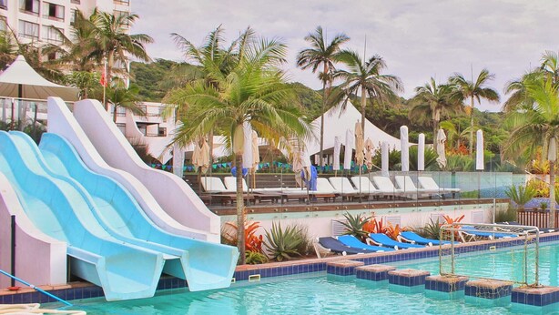 Une piscine avec un toboggan, des chaises longues, des palmiers et un bâtiment d’hôtel en arrière-plan.