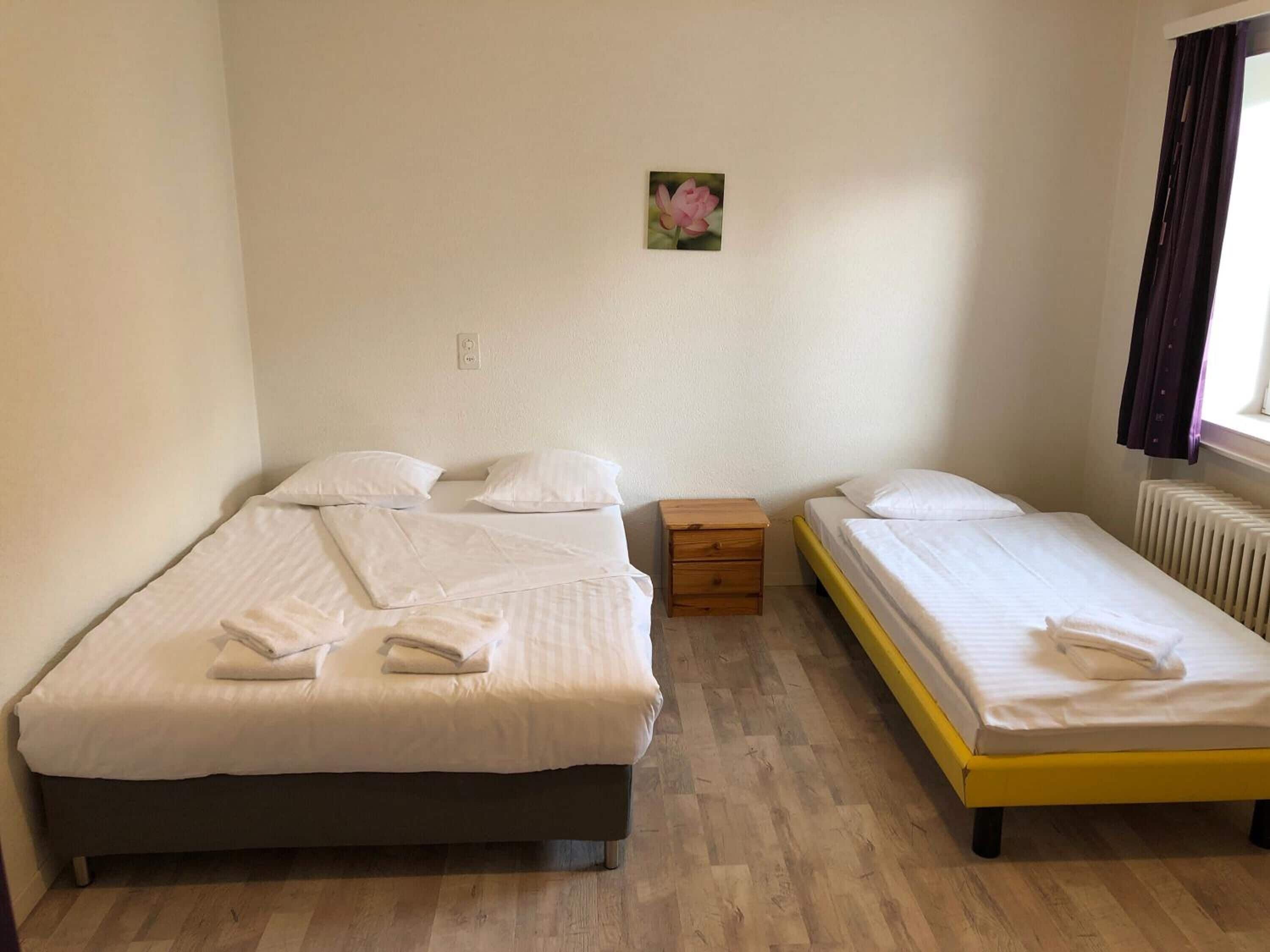 Een kamer met twee bedden, een klein houten nachtkastje, een lichtschakelaar aan de muur en een raam met gordijnen.