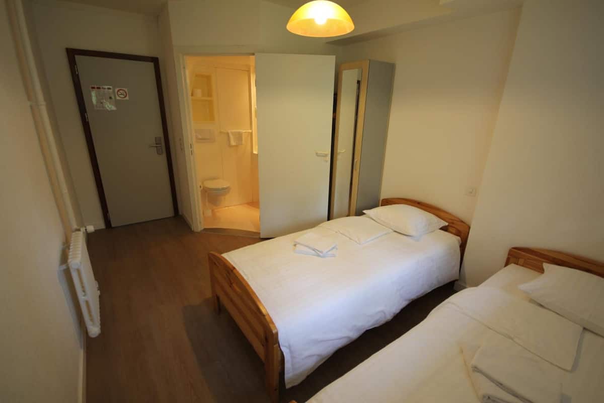 Un dormitorio con dos camas, suelo de madera, un radiador y un baño visible a través de una puerta abierta.