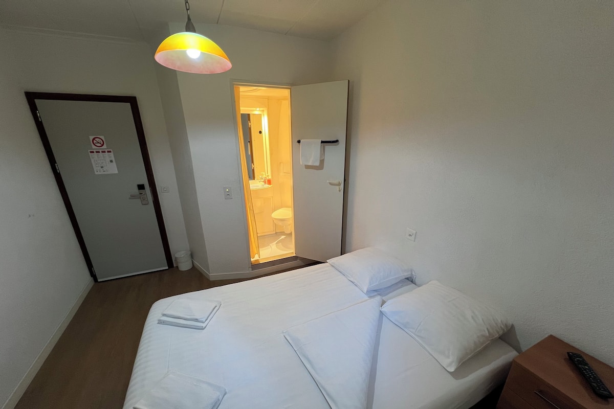 Una cama individual con ropa de cama blanca, una mesita de noche con un control remoto y una puerta que da a un baño.