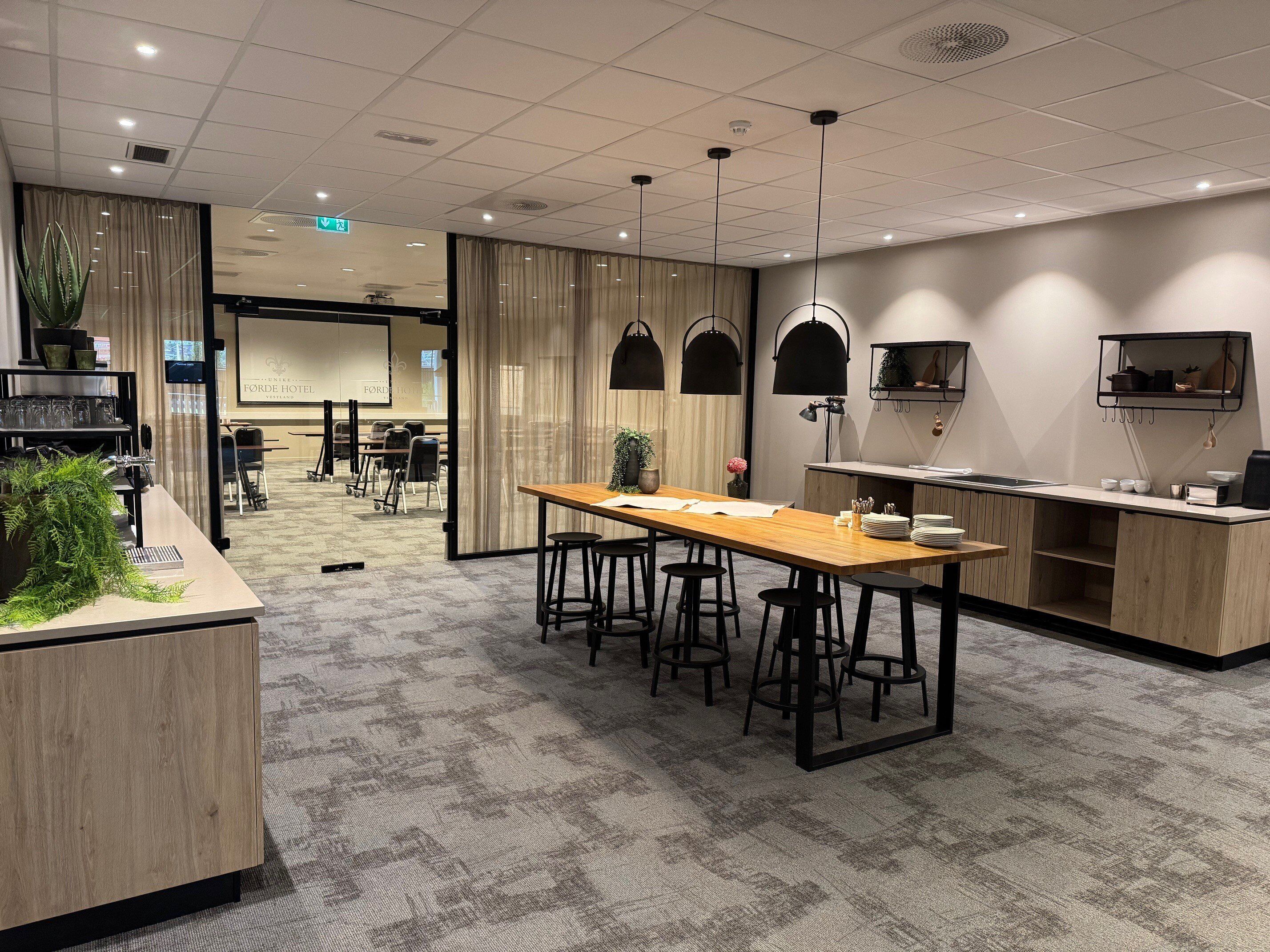 Habitación de hotel moderna con escritorio de madera, taburetes de bar, una zona de cocina y una sala de conferencias al fondo.