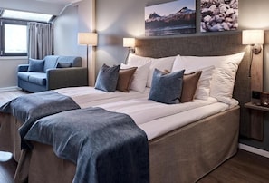 Una cama doble con cabecera, dos mesitas de noche con lámparas, un sillón azul y una ventana con cortinas.