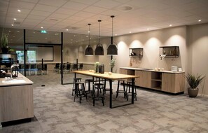 Una sala de conferencias moderna con una mesa larga, varias sillas, una zona de cocina y un ventanal grande.