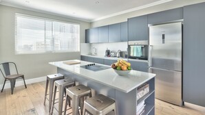 Una cocina moderna con gabinetes grises, un refrigerador de acero inoxidable, una isla central con taburetes y una ventana con persianas.