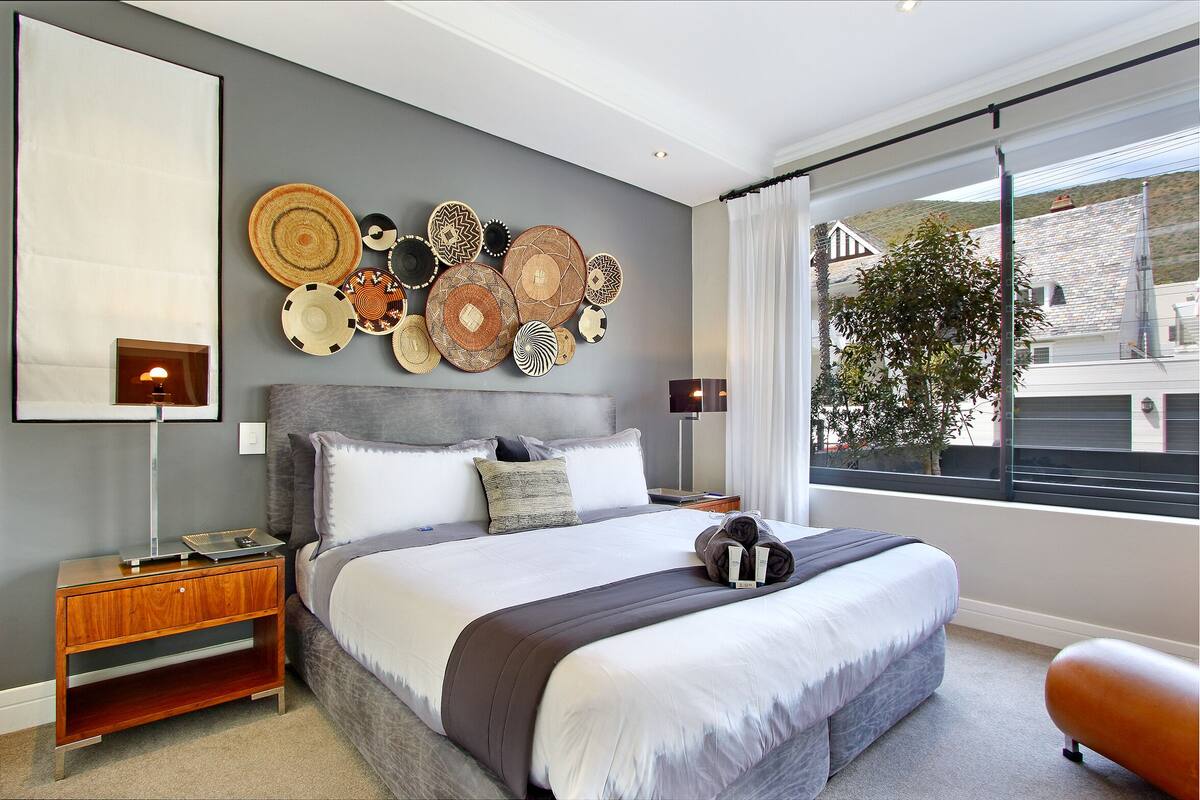 A modern bedroom with a large bed, a nightstand, a wall-mounted lamp, and a view of the outside through a large window.