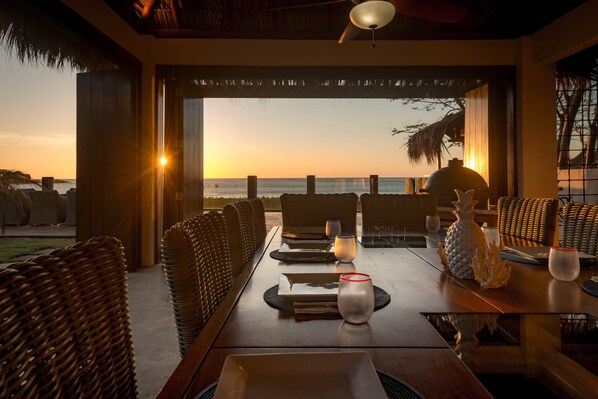 Un comedor con vistas a la puesta de sol sobre el océano, mobiliario de mimbre y una mesa dispuesta con platos, vasos y una piña.