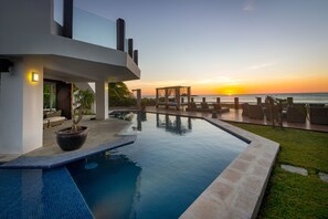 Un área de piscina moderna con vistas a la puesta de sol sobre el océano, una pérgola y mobiliario exterior.